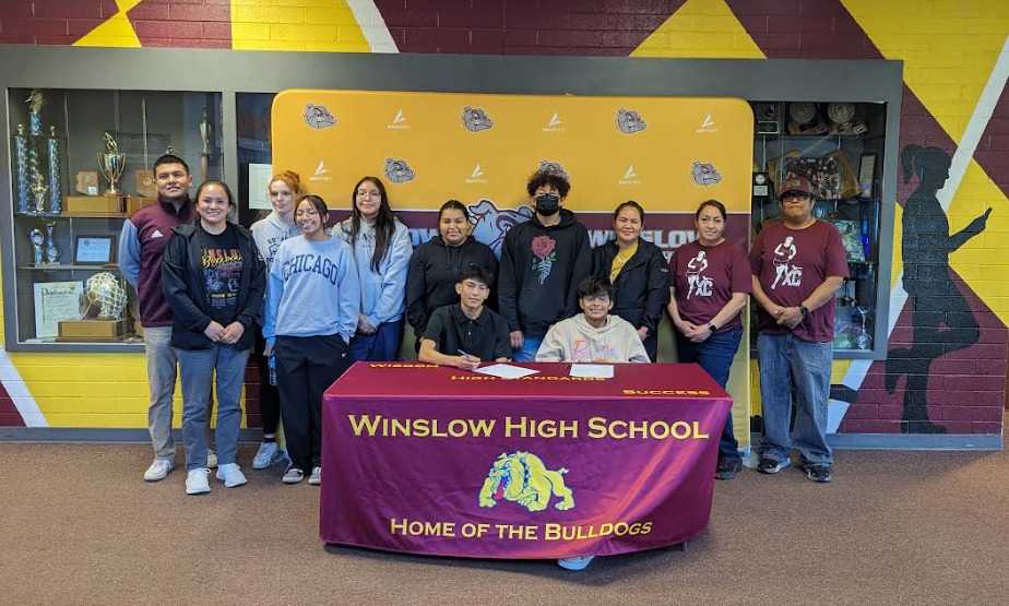 Two athletes sitting at a table with their families standing behind them posing for a photo